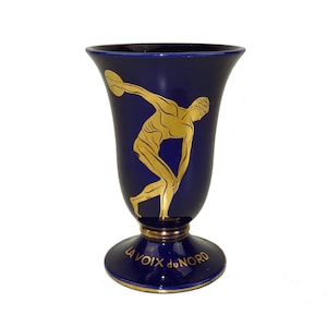 May include: A cobalt blue ceramic vase with a gold-painted image of a discus thrower. The vase has a gold rim and base with the text "LA VOIX du NORD" in gold.