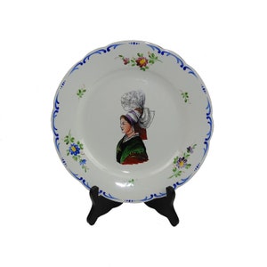 Peut inclure: Une assiette en céramique blanche avec une bordure bleue et blanche. L'assiette présente une image peinte à la main d'une femme en costume traditionnel avec un couvre-chef blanc. La femme regarde vers la gauche et porte une robe verte et rouge. L'assiette est sur un support noir.