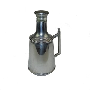 May include: A silver metal dispenser with a handle and a spout. The dispenser has a rounded body and a cylindrical top.