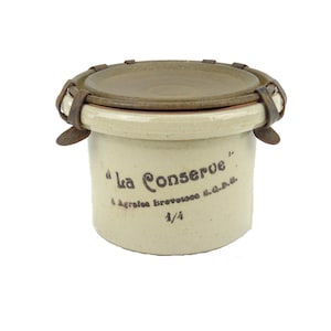 Peut inclure: Un pot en céramique blanche avec un couvercle brun et des fermoirs métalliques. Le pot est étiqueté "La Conserve" et "A. Agricola Brevetata & C.P.R.". Le pot est rempli à 1/4.