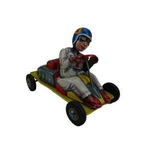 Peut inclure: Une voiture de course jouet jaune et rouge avec une clé de remontage. La voiture a un conducteur portant un casque bleu et une combinaison de course blanche. Le conducteur tient un volant.