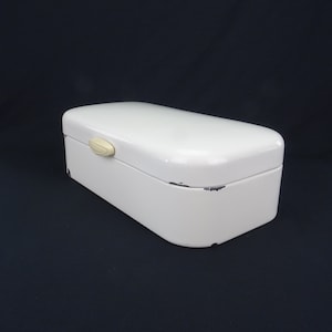 Peut inclure: Boîte à pain en émail blanc avec un dessus légèrement arrondi et une petite poignée blanc cassé. La boîte a un aspect vieilli et vintage avec quelques éclats d'émail.