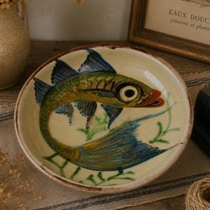 May include: A ceramic plate with a hand-painted fish design. The fish has a green body, blue fins, and a red mouth. The plate has a cream-colored background with green plant accents and a brown rim. The plate is a decorative item.
