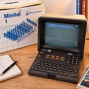 May include: A vintage Minitel 1 terminal with a dark grey keyboard and screen displaying text, accompanied by its original white cardboard box featuring blue branding. The device includes a numeric keypad and function keys, sitting alongside a notebook.