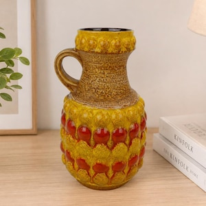 Peut inclure: Un vase en céramique présentant un vernis texturé jaune moutarde avec un motif répétitif de bosses circulaires en relief. La partie inférieure du vase affiche des accents rouge vif dans les creux circulaires, et il possède une anse.
