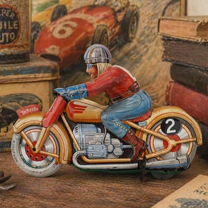 May include: Vintage toy motorcycle with a rider in a red jacket and blue jeans. The motorcycle is yellow and red with the number 2 on the wheel. The word "Technofix" is printed on the side. Other vintage items are in the background.