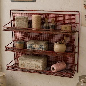May include: A red metal wall-mounted shelf with three tiers of wire mesh shelving. The shelves hold various items including a spool of twine, small glass bottles, a ceramic pot, and rectangular metal tins with floral and text designs.