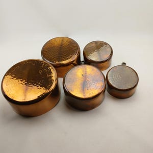 K&ouml;nnte beinhalten: Ein Set aus f&uuml;nf Kupferpfannen mit geh&auml;mmerten Texturen. Die Pfannen variieren in der Gr&ouml;&szlig;e und haben abgerundete K&ouml;rper und Deckel. Das Kupfer hat einen warmen, r&ouml;tlich-braunen Farbton, und die Oberfl&auml;chen reflektieren das Licht. Die Pfannen sind auf einer wei&szlig;en Oberfl&auml;che angeordnet.