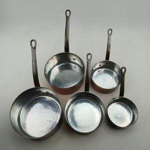 K&ouml;nnte beinhalten: Ein Set aus sechs Kupfer-Kocht&ouml;pfen mit geh&auml;mmerten Innenr&auml;umen und langen, genieteten Griffen. Die Pfannen variieren in der Gr&ouml;&szlig;e und haben einen gl&auml;nzenden Innenraum und ein dunkles, gealtertes &Auml;u&szlig;eres. Die Griffe haben eine Schlaufe zum Aufh&auml;ngen.