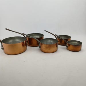 K&ouml;nnte beinhalten: Ein Set aus f&uuml;nf Kupfer-Kocht&ouml;pfen mit langen, silberfarbenen Griffen. Die T&ouml;pfe haben eine gl&auml;nzende Kupferau&szlig;enseite und eine silberfarbene Innenseite. Die Kocht&ouml;pfe sind in verschiedenen Gr&ouml;&szlig;en erh&auml;ltlich, der gr&ouml;&szlig;te misst etwa 20 cm im Durchmesser.