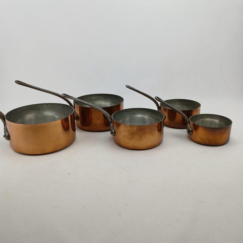 K&ouml;nnte beinhalten: Ein Set aus f&uuml;nf Kupfer-Kocht&ouml;pfen mit langen, silberfarbenen Griffen. Die T&ouml;pfe haben eine gl&auml;nzende Kupferau&szlig;enseite und eine silberfarbene Innenseite. Die Kocht&ouml;pfe sind in verschiedenen Gr&ouml;&szlig;en erh&auml;ltlich, der gr&ouml;&szlig;te misst etwa 20 cm im Durchmesser.