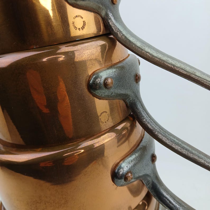 K&ouml;nnte beinhalten: Nahaufnahme von Kupferkochgeschirr mit genieteten Eisengriffen. Die Kupfert&ouml;pfe haben eine gl&auml;nzende, reflektierende Oberfl&auml;che. Die Griffe sind mit Nieten befestigt. Der Text "Atelier & Calvet" ist auf das Kupfer gestempelt.