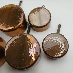 K&ouml;nnte beinhalten: Eine Sammlung von Kupfer-T&ouml;pfen und -Pfannen mit geh&auml;mmerten Oberfl&auml;chen. Das Kochgeschirr umfasst verschiedene Gr&ouml;&szlig;en, jeweils mit einem Griff und einem gl&auml;nzenden, reflektierenden Finish. Die Kupferfarbe ist ein warmes, r&ouml;tliches Braun.