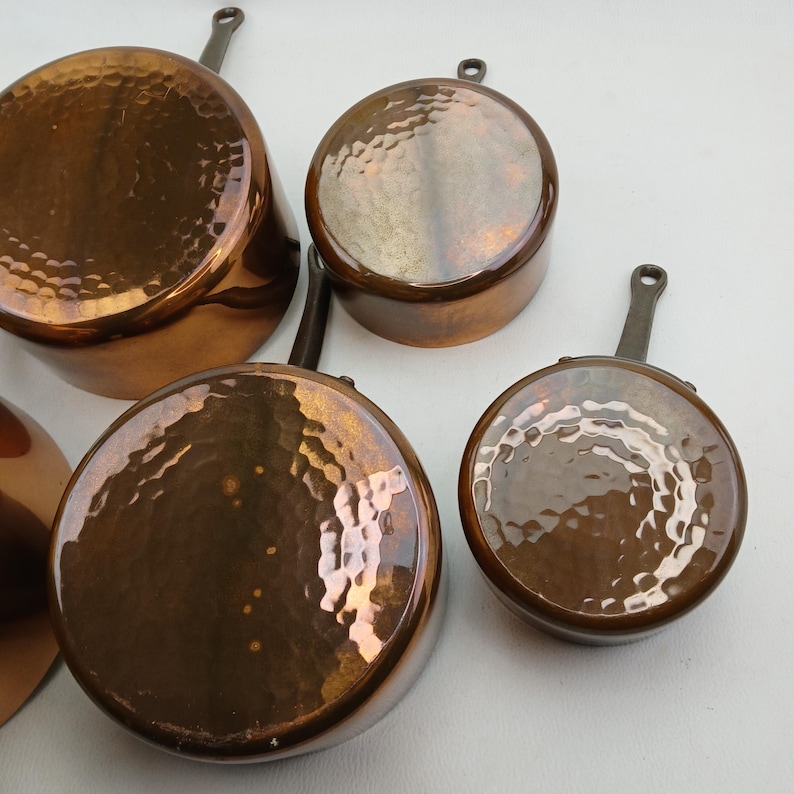 K&ouml;nnte beinhalten: Eine Sammlung von Kupfer-T&ouml;pfen und -Pfannen mit geh&auml;mmerten Oberfl&auml;chen. Das Kochgeschirr umfasst verschiedene Gr&ouml;&szlig;en, jeweils mit einem Griff und einem gl&auml;nzenden, reflektierenden Finish. Die Kupferfarbe ist ein warmes, r&ouml;tliches Braun.