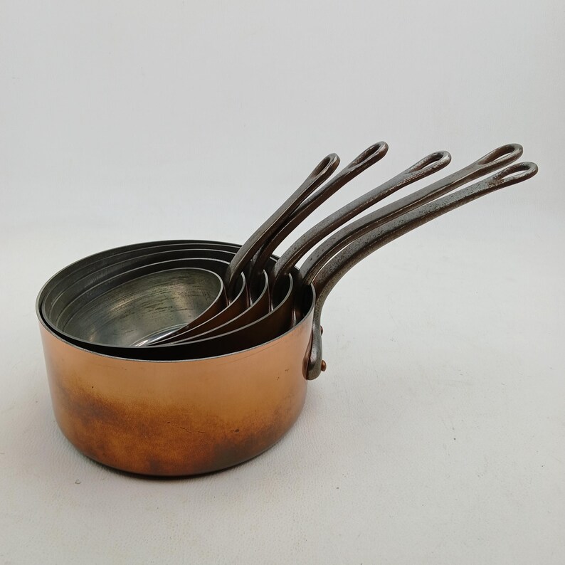 K&ouml;nnte beinhalten: Ein Satz von f&uuml;nf Kupfer-Kocht&ouml;pfen mit genieteten, dunklen Metallgriffen. Die T&ouml;pfe sind ineinander verschachtelt, wobei sich der gr&ouml;&szlig;te im Vordergrund befindet. Das Kupfer&auml;u&szlig;ere hat einen warmen, r&ouml;tlich-braunen Farbton.