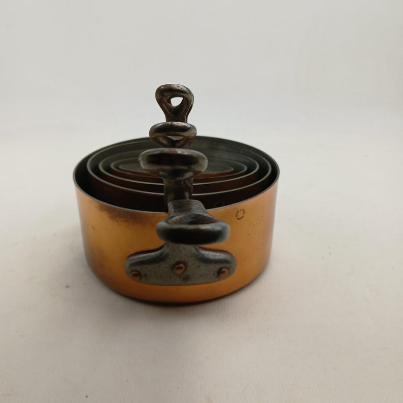 K&ouml;nnte beinhalten: Ein Satz kupferfarbener, runder Ausstecher in verschiedenen Gr&ouml;&szlig;en, die ineinander verschachtelt sind. Ein Metallgriff mit einer Schlaufe und dekorativen Elementen ist an dem gr&ouml;&szlig;ten Ausstecher befestigt. Die Ausstecher befinden sich auf einer wei&szlig;en Oberfl&auml;che.