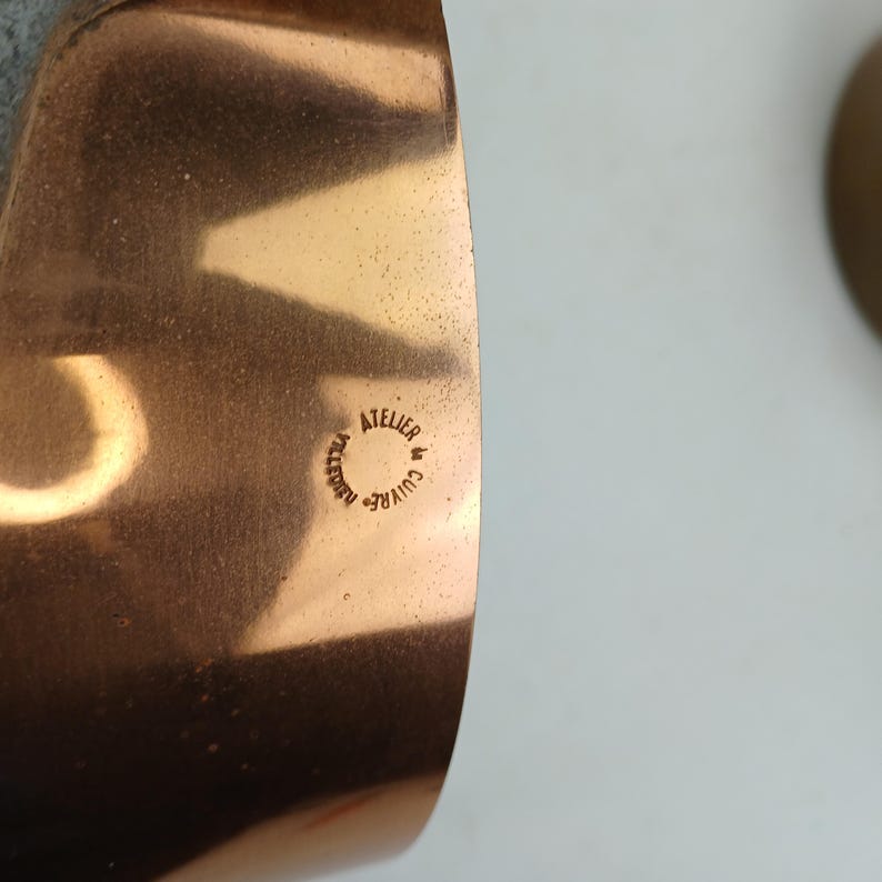 K&ouml;nnte beinhalten: Nahaufnahme eines Kupferarmbands mit poliertem Finish. Das Armband hat ein geschwungenes Design und tr&auml;gt den Text "ATELIER CUIVRE" in einem kreisf&ouml;rmigen Stempel. Das Kupfer hat einen warmen, r&ouml;tlich-braunen Farbton.