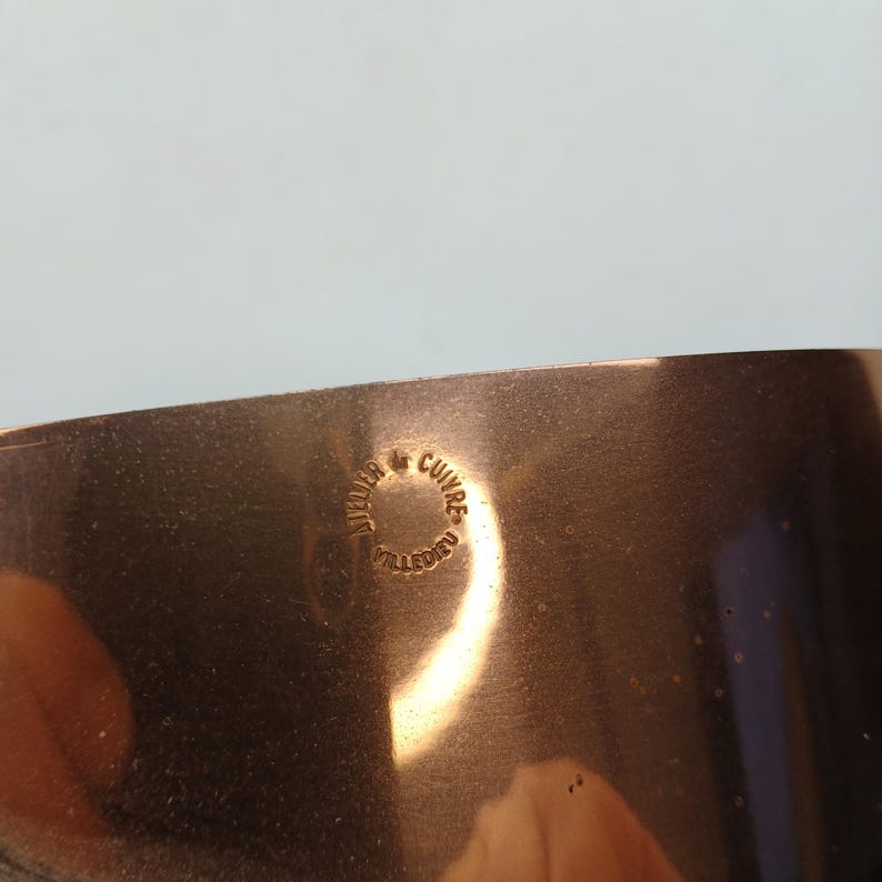 K&ouml;nnte beinhalten: Nahaufnahme eines Kupfertopfes mit gl&auml;nzender, reflektierender Oberfl&auml;che. Der Topf hat eine runde Form und eine glatte Textur. Ein kreisf&ouml;rmiger Stempel mit dem Text "Atelier & Cuivres Villedieu" ist sichtbar.