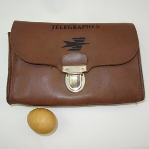Może przedstawiać: Brązowa sk&oacute;rzana torebka z srebrnym zapięciem i napisem "TELEGRAPHES" oraz stylizowanym logo ptaka.