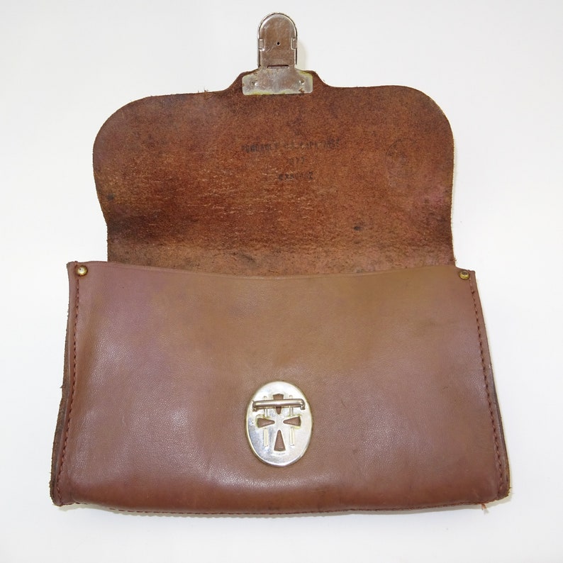 Może przedstawiać: Brązowa sk&oacute;rzana torebka z srebrnym zapięciem i srebrnym wzorem w kształcie krzyża z przodu. Torebka jest otwarta i widać wnętrze.