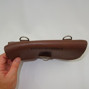 Może przedstawiać: Brązowa sk&oacute;rzana torebka z srebrnym zapięciem i dwoma metalowymi k&oacute;łkami. Torebka jest oznaczona napisem "TELEGRAPHES".