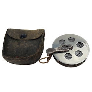 Könnte beinhalten: Ein antikes Vermessungswerkzeug-Set, bestehend aus einem Metallmessrad mit Richtungsanzeiger und einer Ledertasche. Das Rad ist silberfarben, und die Tasche ist dunkelbraun mit einem schwarzen Knopfverschluss. Ein Messingring ist befestigt.