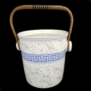 Puede incluir: Un cubo de hielo de cerámica blanca con un patrón de grecas azules y un asa tejida. El cubo tiene una textura similar al mármol y un borde con dibujos azules. El asa es de ratán tejido.