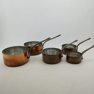 K&ouml;nnte beinhalten: Ein Set aus f&uuml;nf Kupfer-Kocht&ouml;pfen mit langen Griffen. Die T&ouml;pfe sind unterschiedlich gro&szlig;, mit einer gl&auml;nzenden Kupferau&szlig;enseite und einer silberfarbenen Innenseite. Die Griffe sind dunkler, m&ouml;glicherweise aus Bronze.