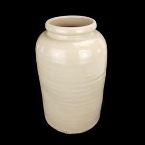 May include: Off-white ceramic jar with a wide opening and a slightly textured surface. The jar has a cylindrical shape with a rounded base and a smooth, rounded rim. The jar is likely used for storage or decorative purposes.