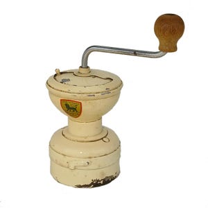May include: A vintage, off-white manual coffee grinder with a wooden handle. The grinder has a lion logo and the text "Made in France" on the side.
