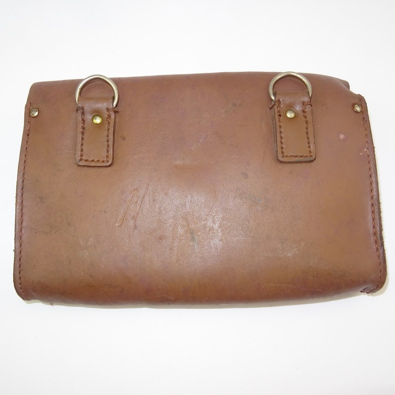 Może przedstawiać: Brązowa sk&oacute;rzana torba listonoszka z dwoma srebrnymi klamrami i klapką. Torba ma zużyty wygląd i jest wykonana z wytrzymałego materiału.