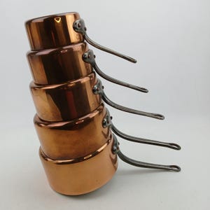 K&ouml;nnte beinhalten: Ein Stapel von f&uuml;nf Kupfer-Kocht&ouml;pfen mit dunklen Metallgriffen. Die T&ouml;pfe sind in absteigender Reihenfolge nach Gr&ouml;&szlig;e angeordnet, wobei der gr&ouml;&szlig;te Topf unten ist. Das Kupfer hat eine warme, reflektierende Oberfl&auml;che, und die Griffe sind an den Seiten vernietet.