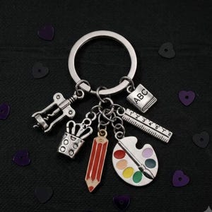May include: A silver-toned metal keyring featuring five dangling charms: a corkscrew, a pencil holder, a red pencil, a ruler with markings, and an artist palette with colourful paint spots, plus a small square charm embossed with the letters ABC.