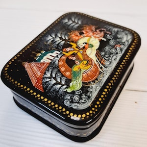 Könnte beinhalten: Eine rechteckige, schwarz lackierte Schatulle mit einer detaillierten, farbenfrohen Malerei auf dem Deckel. Das Kunstwerk zeigt Figuren in traditioneller Kleidung vor dem Hintergrund wirbelnder Muster und architektonischer Elemente. Goldene Punkte umranden den Deckel.