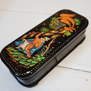 Peut inclure: Une boîte laquée noire de forme rectangulaire aux bords arrondis. La boîte présente une peinture colorée et détaillée d'un personnage vêtu d'orange et de bleu, avec un oiseau doré et du feuillage. La boîte est ornée de points dorés.