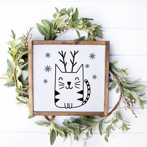 May include: A black and white illustration of a cat with antlers, surrounded by snowflakes. The cat is sitting on a white background inside a wooden frame.