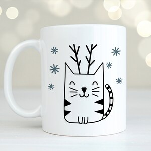 May include: White ceramic mug featuring a black and white illustration of a cat wearing antlers. The cat is surrounded by small snowflakes.