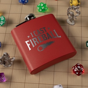 May include: A red flask with the text "I CAST FIREBALL" in silver. The flask is rectangular with a silver cap. The image includes various dice and miniature figurines, suggesting a tabletop gaming theme.