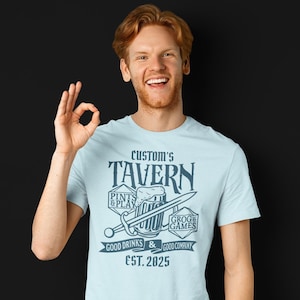 May include: A light blue t-shirt with a vintage tavern logo featuring a beer mug, sword, dice, and the text "Custom's Tavern, Pints & Play, Grog & Games, Good Drinks & Good Company, Est. 2025."