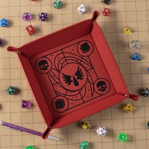 May include: A red, square dice tray with a black graphic design featuring skulls, wings, and geometric shapes. The tray is surrounded by various colorful dice, including D20s, D12s, and D6s. A purple pen rests near the tray.