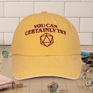 May include: A yellow baseball cap with the text "You can certainly try" and a red 20-sided die embroidered on the front.