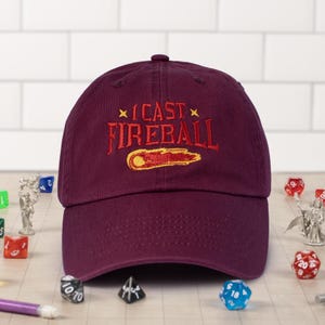 May include: Burgundy baseball cap with an embroidered design that says "I cast fireball" with a yellow and red fireball graphic.