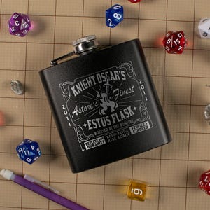 May include: Black flask with silver accents and the text "Knight Oscar's Finest Estus Flask" in white. The flask is surrounded by dice and a purple pencil on a grid-patterned surface. The flask is approximately 4 inches tall.