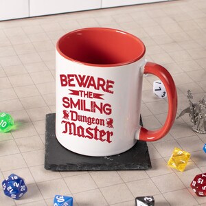 May include: A white ceramic mug with a red interior and handle, featuring the text "BEWARE THE SMILING Dungeon Master" in red. The mug sits on a black square coaster, surrounded by dice, suggesting a tabletop gaming theme.