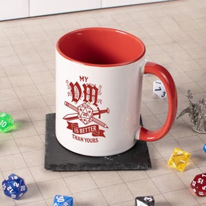 May include: White ceramic mug with a red interior and handle. The mug features the text "MY DM IS BETTER THAN YOURS" in red, along with a graphic of a die, swords, and a banner. The mug sits on a black square coaster.