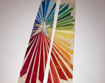 May include: A beige and multicolored liturgical stole with a starburst design. The starburst radiates from a central white star, with sections of red, orange, yellow, green, and blue. The stole is made of fabric with a subtle pattern.