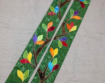 May include: Two green fabric stoles with a tree design. The trees have brown branches and colorful leaves in shades of red, orange, yellow, blue, and purple. The stoles are on a gray background.