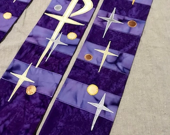 May include: Purple liturgical stole with embroidered symbols. The stole features a dark purple batik fabric with lighter purple stripes and embroidered crosses, stars, and a Chi Rho symbol. Gold and silver accents add detail to the design.