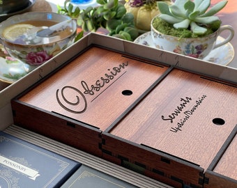 Könnte beinhalten: Eine Holzkiste mit Fächern, mit graviertem Text auf den Deckeln. Der linke Deckel trägt die Aufschrift "Obsession" und der rechte Deckel "Servants Upstairs Downstairs". Die Kiste ist von Teetassen und Pflanzen umgeben.