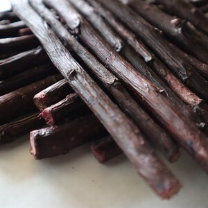 May include: A close-up of a bundle of dark brown sticks, some of which are slightly reddish in color. The sticks are arranged in a criss-cross pattern.